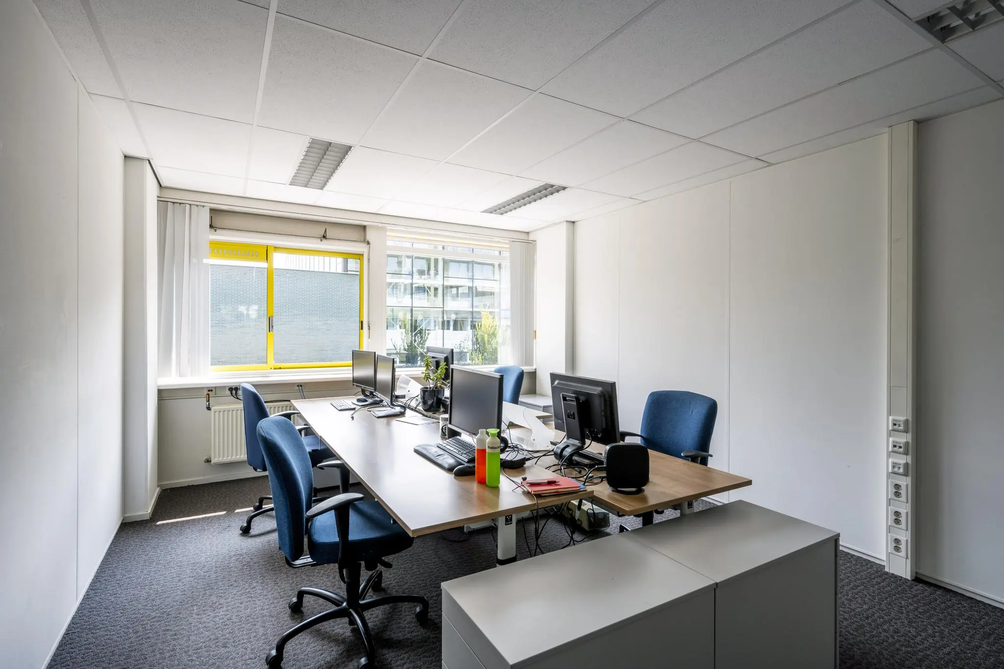 Kantoorruimte aan de Jan Tooropstraat met meerdere bureaus, computerschermen en blauwe bureaustoelen bij een raam met gele kozijnen.